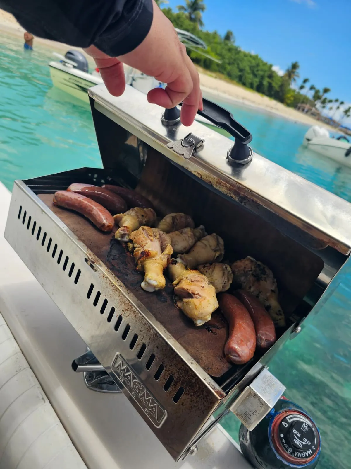 Chicken legs and sausages cooking on the onboard Magma marine grill with turquoise Caribbean water and beach in the background