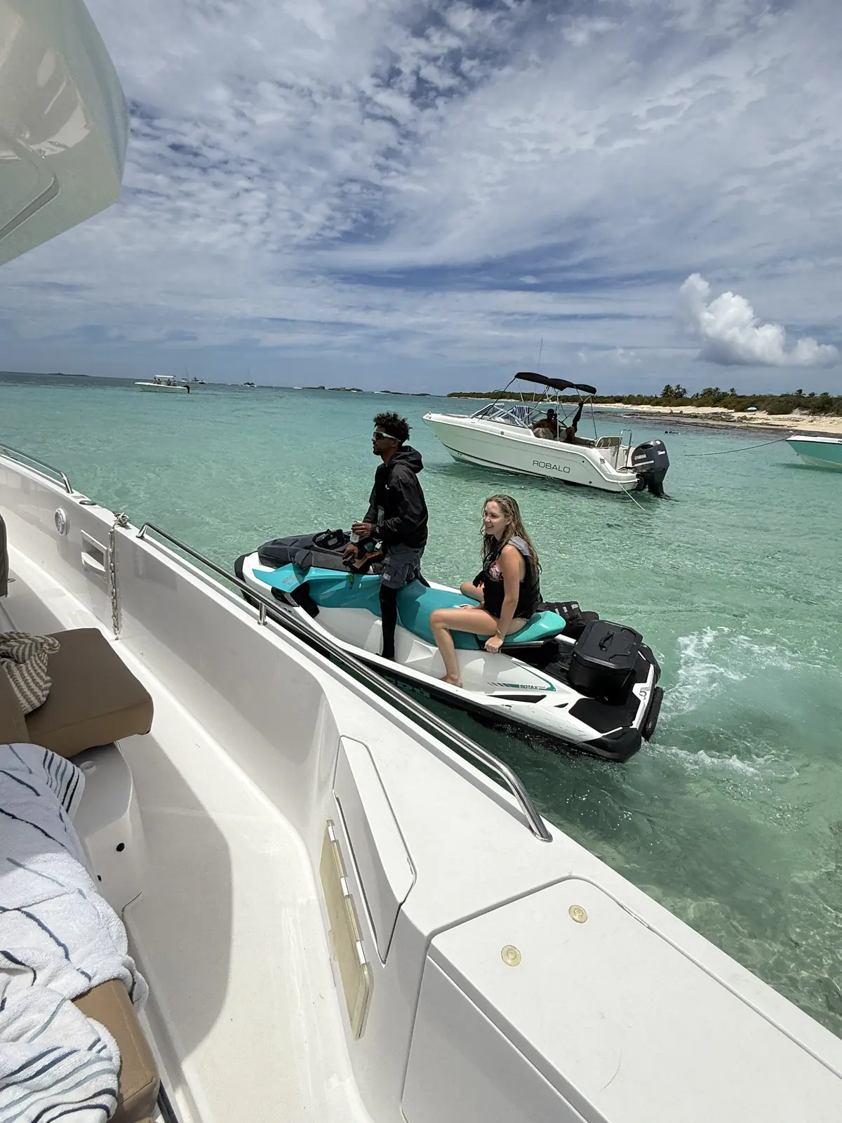 Jet ski tied to yacht with guests, sandbar in background